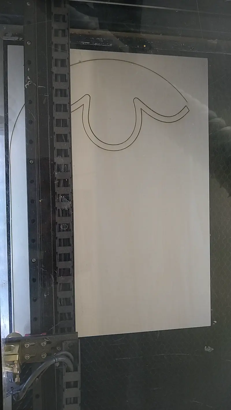 Laser cutting machine engraving a curvilinear design onto a large sheet of plywood, visible through a protective glass cover.