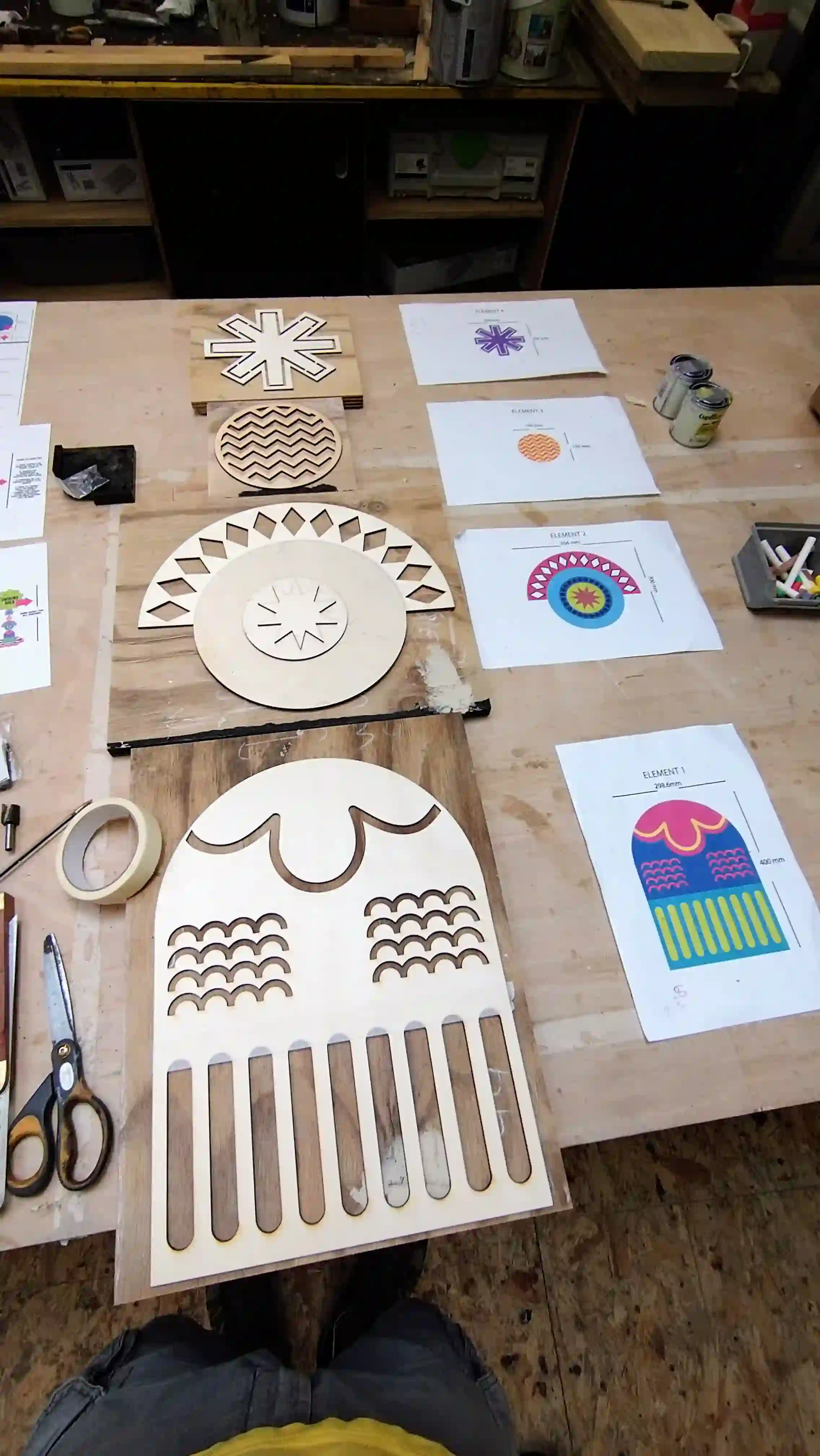 Workshop table displaying laser-cut wooden signage components with geometric and decorative patterns, surrounded by design printouts, scissors, masking tape, and cans of paint.