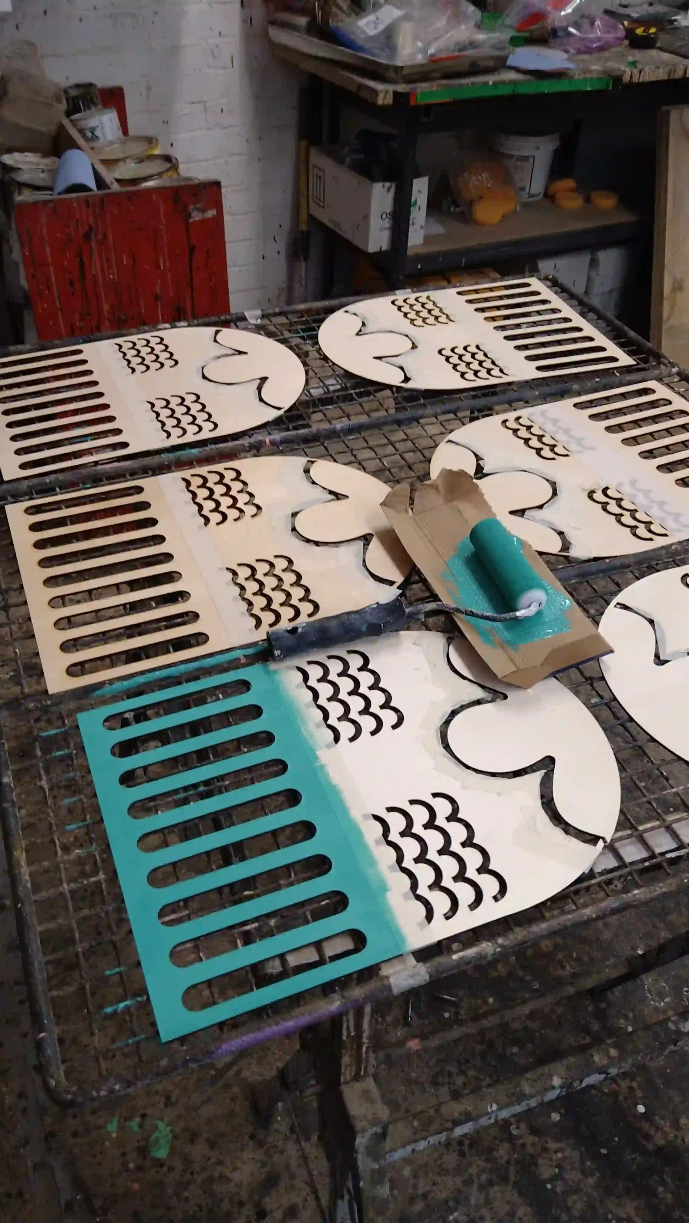 Laser-cut wooden signage pieces laid on a metal grate for painting, with teal paint applied to the lower half using a roller. The workshop space is visible with tools and materials in the background.