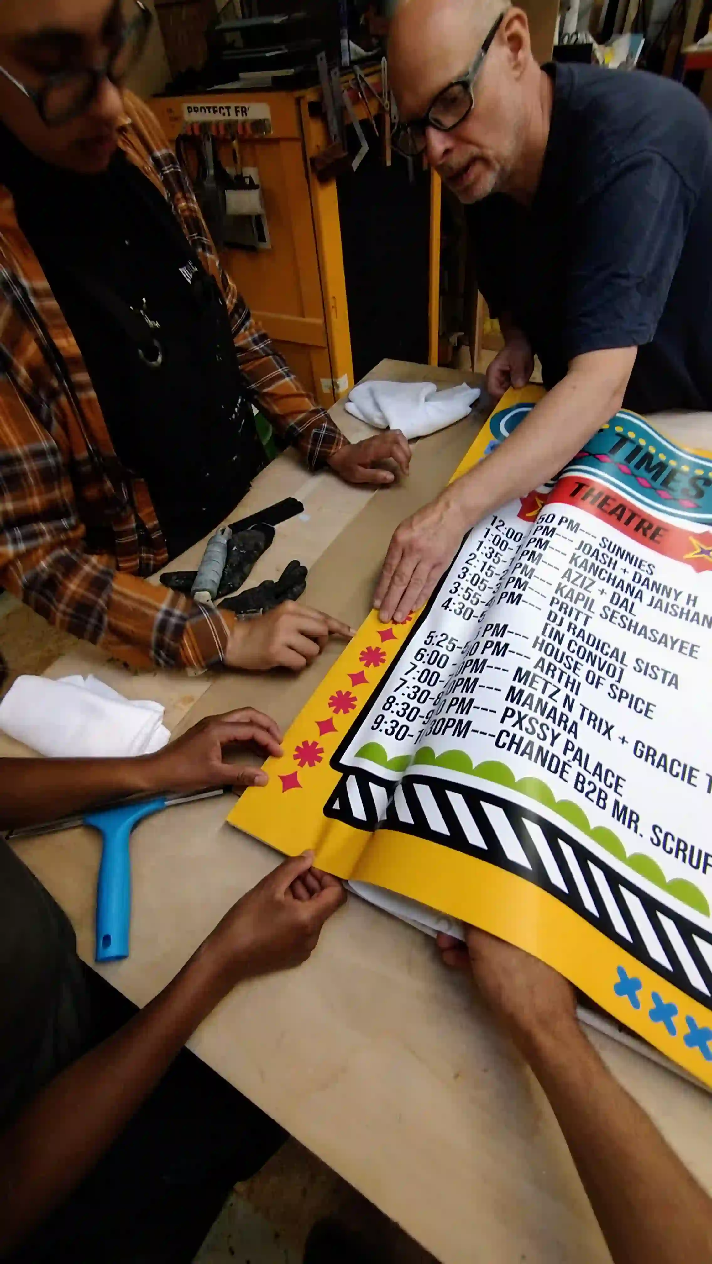 Several hands working together to paste a poster onto a large piece of wooden board.